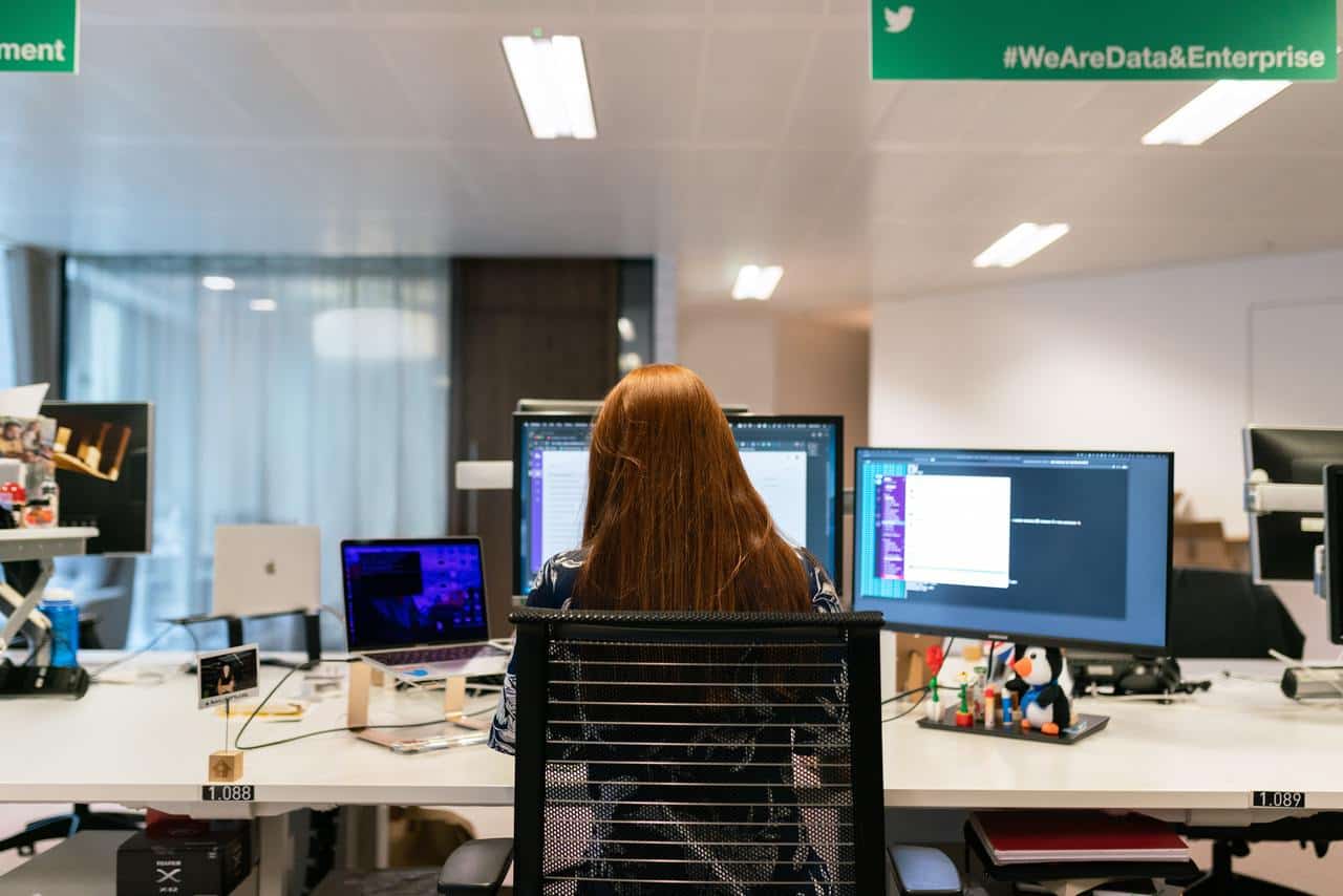 Рыжеволосая женщина работает за компьютером. Перед ней два монитора и ноутбук.